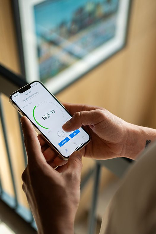 Person holder smarttelefon med app som viser temperaturinnstilling på 19,5 °C i et moderne hjem