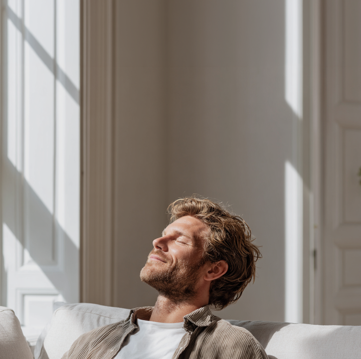 Kvinne sitter avslappet i sofa ved stort vindu i lys stue med lukkede øyne og rolig uttrykk.