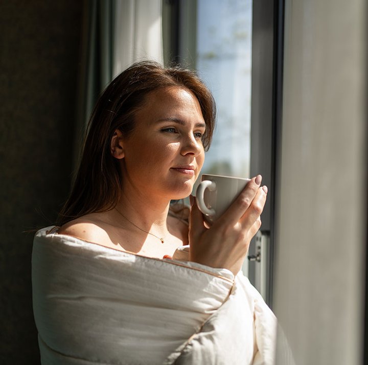 Person står vid ett fönster i bostaden och dricker kaffe i dagsljus, med lugn morgonstämning och naturligt ljus.