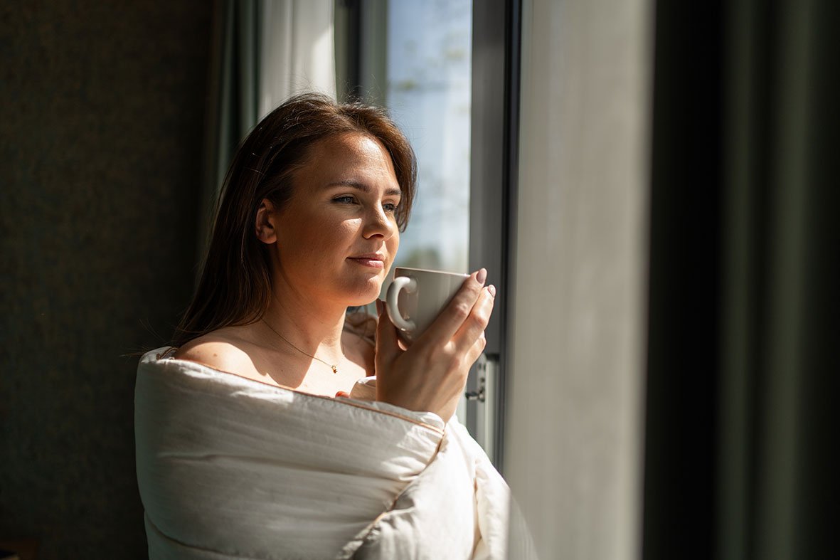 Person står ved vindu i bolig og drikker kaffe i dagslys, med rolig morgenstemning og naturlig lys.