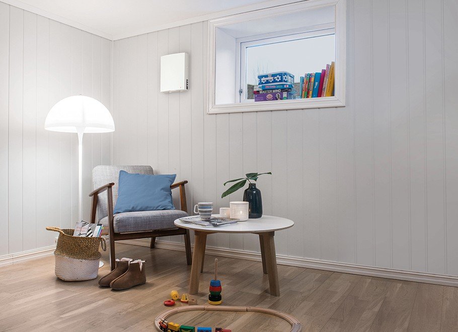 Romventilator montert på vegg i innredet kjellerrom med sittegruppe og leker på gulvet.