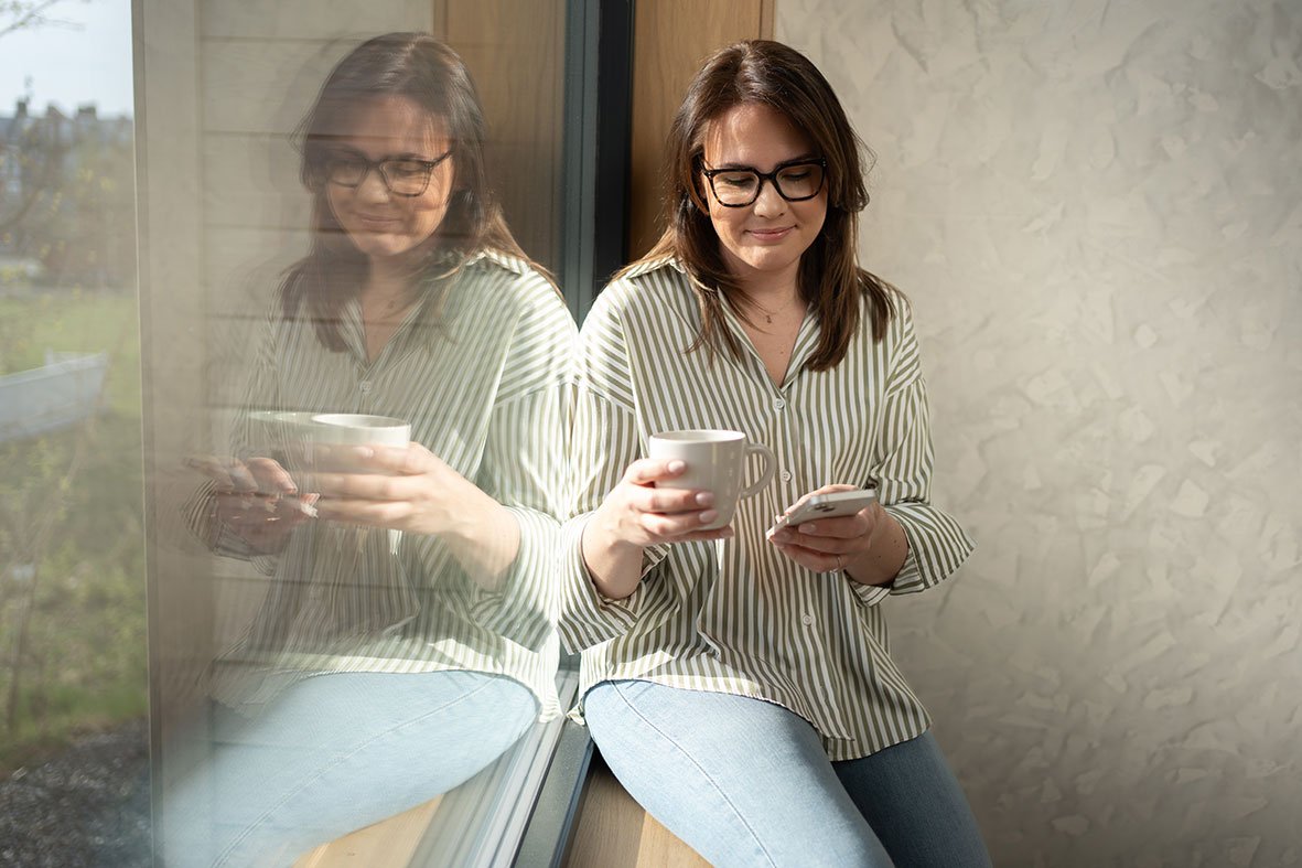 Person using the Flexit GO app on a mobile phone to control ventilation and indoor climate at home, while sitting by a window.