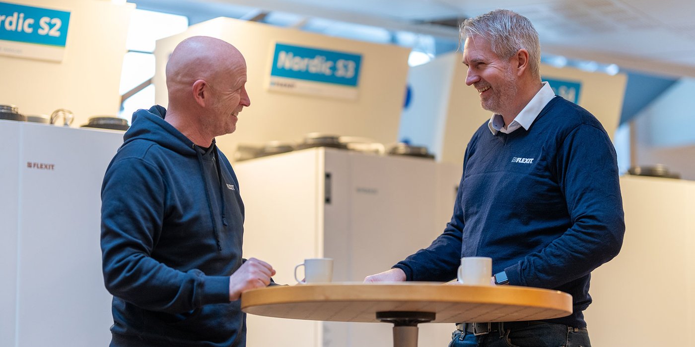 To medarbeidere i Flexits lokaler samtaler ved ståbord, med ventilasjonsaggregater utstilt i bakgrunnen.