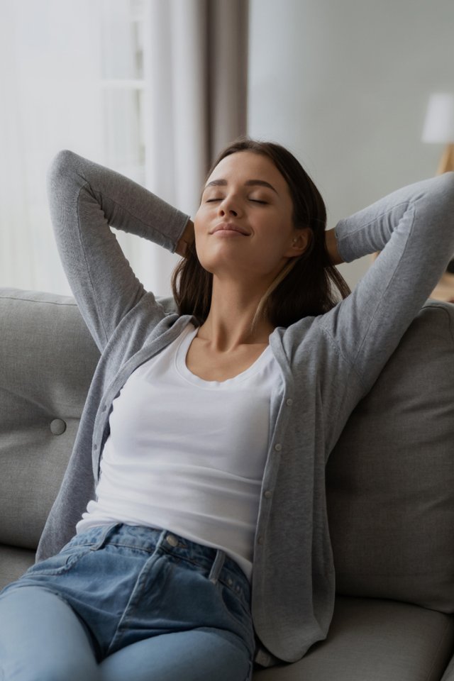 Kvinne sitter avslappet i sofa ved stort vindu i lys stue med lukkede øyne og rolig uttrykk.