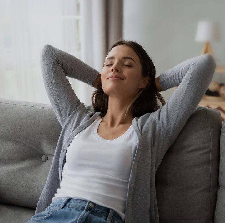 Kvinne sitter avslappet i sofa ved stort vindu i lys stue med lukkede øyne og rolig uttrykk.