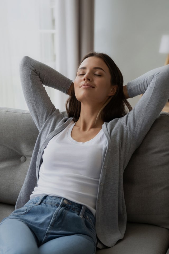 Kvinne sitter avslappet i sofa ved stort vindu i lys stue med lukkede øyne og rolig uttrykk.